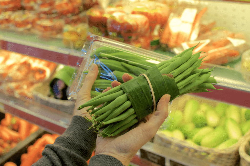Close-up view of a person’s hands holding green beans in a grocery store, one bundle wrapped in banana leaves, the other in a plastic clamshell container.