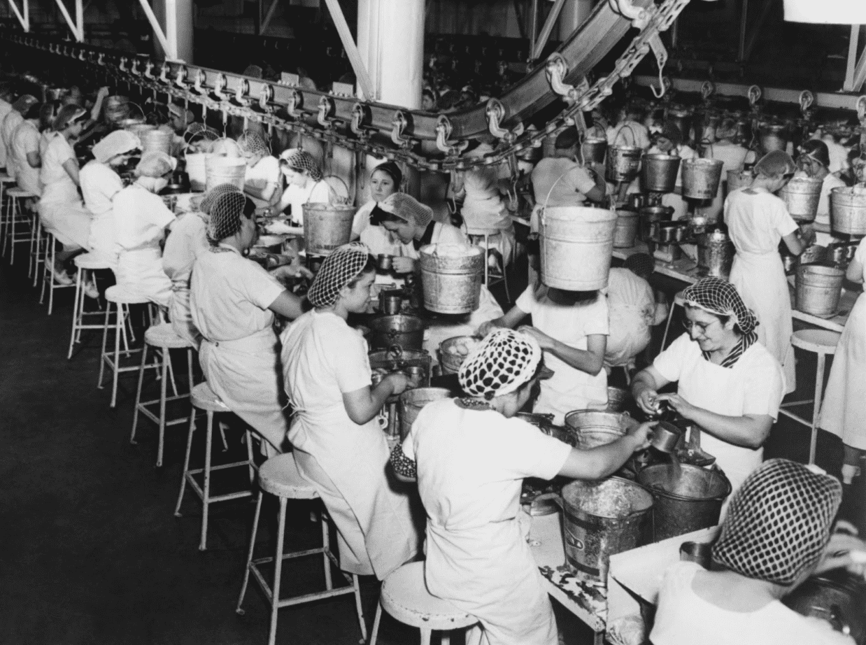 Greyscale photo of an old factory with rows of workers on metal stools hand-processing goods.